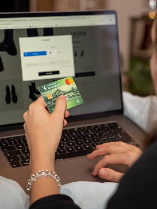 Eine Frau sitzt vor einem Laptop und hat eine Shoppingseite geöffnet. in der linken Hand hält sie ihre Kreditkarte, während sie mit der rechten tippt. Eine Frau sitzt vor einem Laptop und hat eine Shoppingseite geöffnet. in der linken Hand hält sie ihre Kreditkarte, während sie mit der rechten tippt.