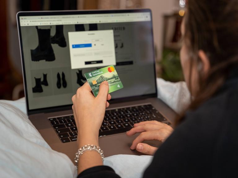 Eine Frau sitzt vor einem Laptop und hat eine Shoppingseite geöffnet. in der linken Hand hält sie ihre Kreditkarte, während sie mit der rechten tippt. Eine Frau sitzt vor einem Laptop und hat eine Shoppingseite geöffnet. in der linken Hand hält sie ihre Kreditkarte, während sie mit der rechten tippt.