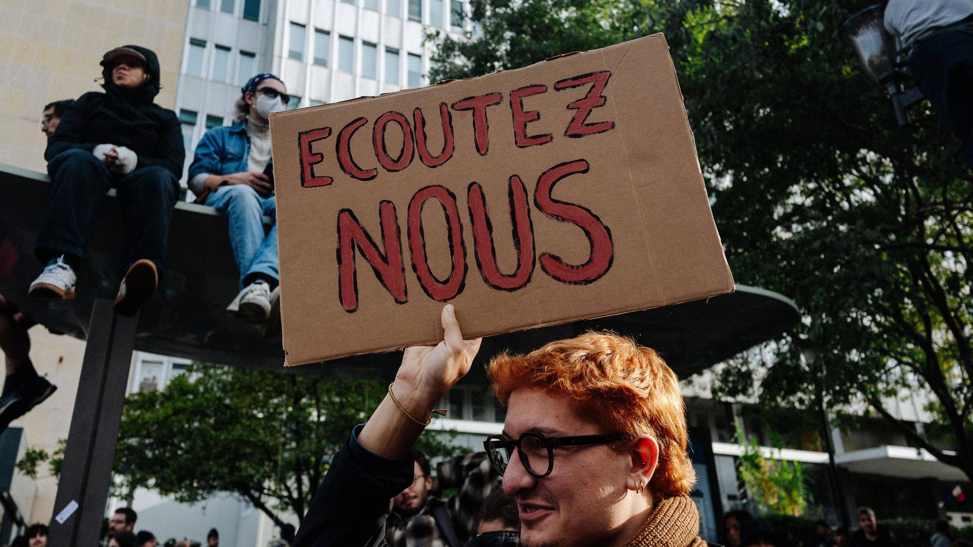 Ein junger Mann mit Brille und Schal hält bei einer Demonstration in Paris ein Pappschild mit der Aufschrift „ECOUTEZ NOUS“ (Hört uns zu) in die Höhe. Im Hintergrund sitzen weitere Protestierende auf einem Unterstand, während im unteren Bildbereich eine… Ein junger Mann mit Brille und Schal hält bei einer Demonstration in Paris ein Pappschild mit der Aufschrift „ECOUTEZ NOUS“ (Hört uns zu) in die Höhe. Im Hintergrund sitzen weitere Protestierende auf einem Unterstand, während im unteren Bildbereich eine…