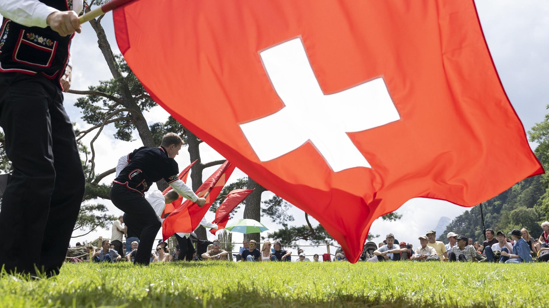 Auf der Rütliwiese in der Schweiz stehen Fahnenschwinger in traditioneller Kleidung; sie schwingen Schweizer-Flaggen (ein weißes Kreuz auf rotem Grund). Auf der Wiese sitzen Menschen und schauen zu.