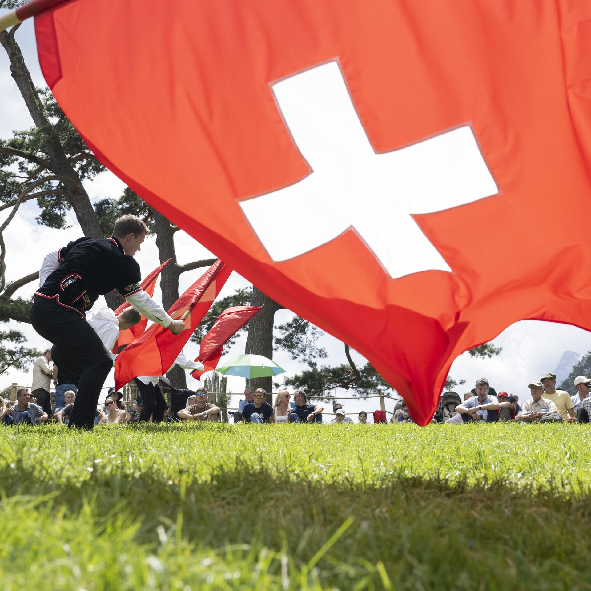 Auf der Rütliwiese in der Schweiz stehen Fahnenschwinger in traditioneller Kleidung; sie schwingen Schweizer-Flaggen (ein weißes Kreuz auf rotem Grund). Auf der Wiese sitzen Menschen und schauen zu. Es ist die Bundesfeier am 1. August 2025; Foto: dpa.