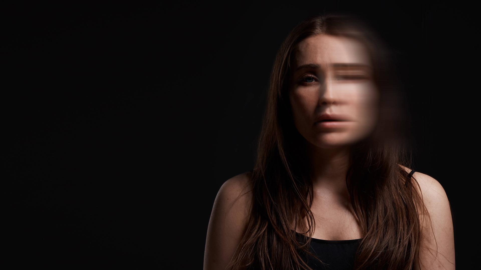 Eine junge Frau mit langen schwarzen Haaren steht vor einem schwarzen Hintergrund. Durch einen Foto-Effekt wirkt eine Gesichtshälfte verschwommen.