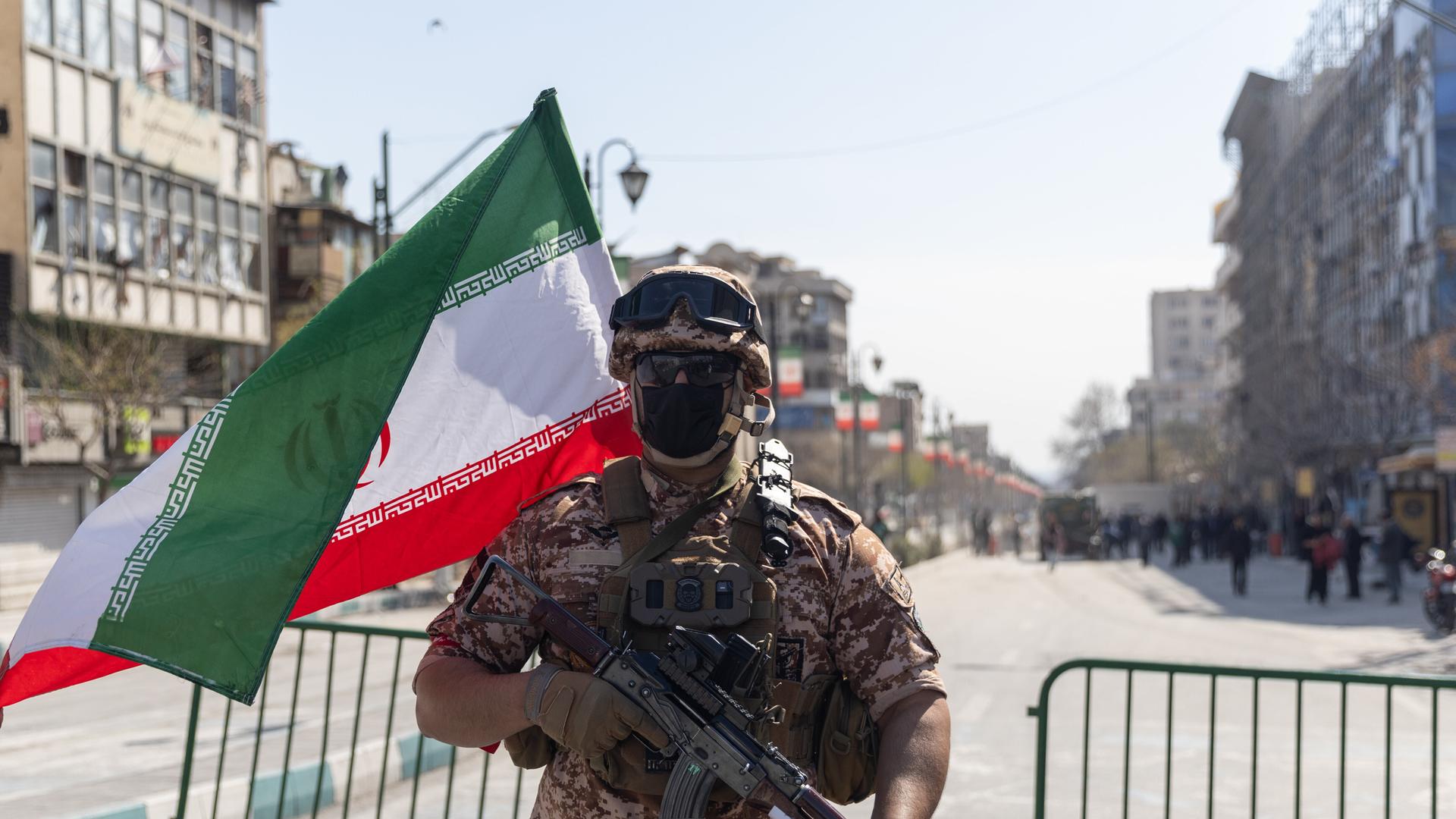 Ein Soldat steht in Iran auf einer Straße und hält ein Maschinengewehr. Hinter ihm steht ein Gitter, eine Iran-Flagge weht im Wind.