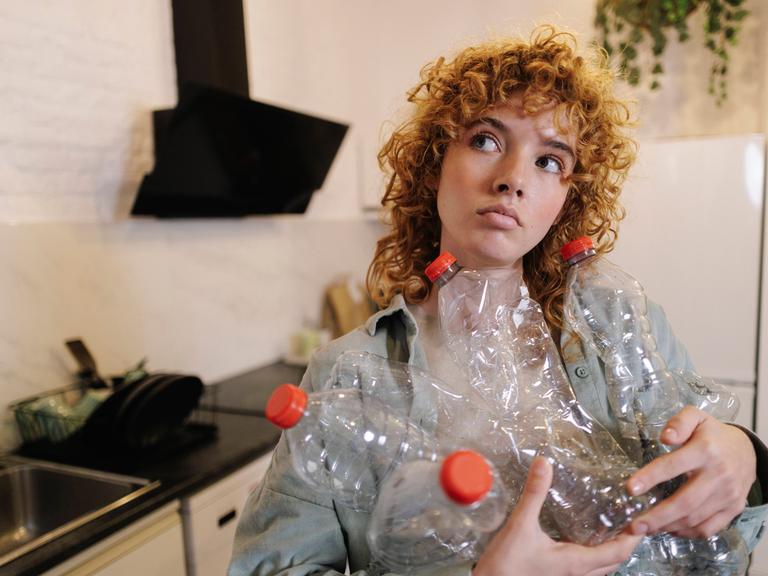 Eine junge Frau steht in einer Küche und hält viele leere Pfandflaschen in den Händen.