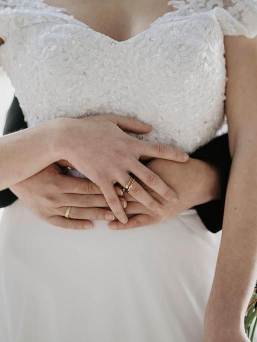 Ein Hochzeitspaar steht hintereinander. Eine der beiden Frauen trägt ein weißes Hochzeitskleid. Die andere Frau trägt einen schwarzen Anzug. Sie umarmen sich, dabei halten sie ihre Hände übereinander. Ein Hochzeitspaar steht hintereinander. Eine der beiden Frauen trägt ein weißes Hochzeitskleid. Die andere Frau trägt einen schwarzen Anzug. Sie umarmen sich, dabei halten sie ihre Hände übereinander.