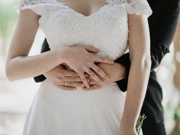 Ein Hochzeitspaar steht hintereinander. Eine der beiden Frauen trägt ein weißes Hochzeitskleid. Die andere Frau trägt einen schwarzen Anzug. Sie umarmen sich, dabei halten sie ihre Hände übereinander. Ein Hochzeitspaar steht hintereinander. Eine der beiden Frauen trägt ein weißes Hochzeitskleid. Die andere Frau trägt einen schwarzen Anzug. Sie umarmen sich, dabei halten sie ihre Hände übereinander.