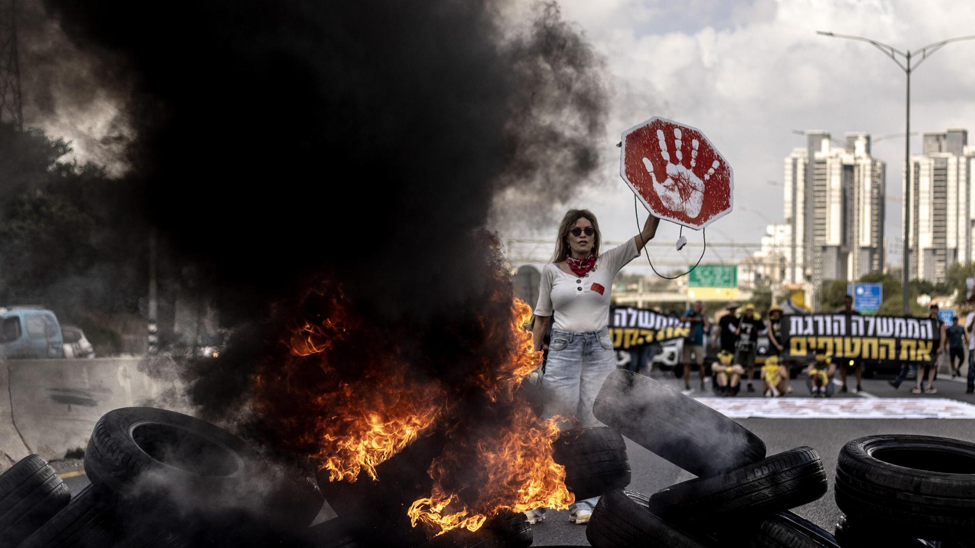 Demonstrantin in Tel Aviv (Israel) hält ein rotes Schild mit weißem Handabdruck in die Höhe; vor ihr liegt eine Barrikade aus brennenden Reifen, neben ihr steigt eine schwarze Rauchwolke in die Höhe (26.08.2025) Demonstrantin in Tel Aviv (Israel) hält ein rotes Schild mit weißem Handabdruck in die Höhe; vor ihr liegt eine Barrikade aus brennenden Reifen, neben ihr steigt eine schwarze Rauchwolke in die Höhe (26.08.2025)