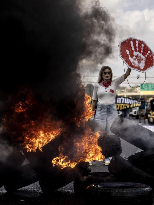 Demonstrantin in Tel Aviv (Israel) hält ein rotes Schild mit weißem Handabdruck in die Höhe; vor ihr liegt eine Barrikade aus brennenden Reifen, neben ihr steigt eine schwarze Rauchwolke in die Höhe (26.08.2025)
