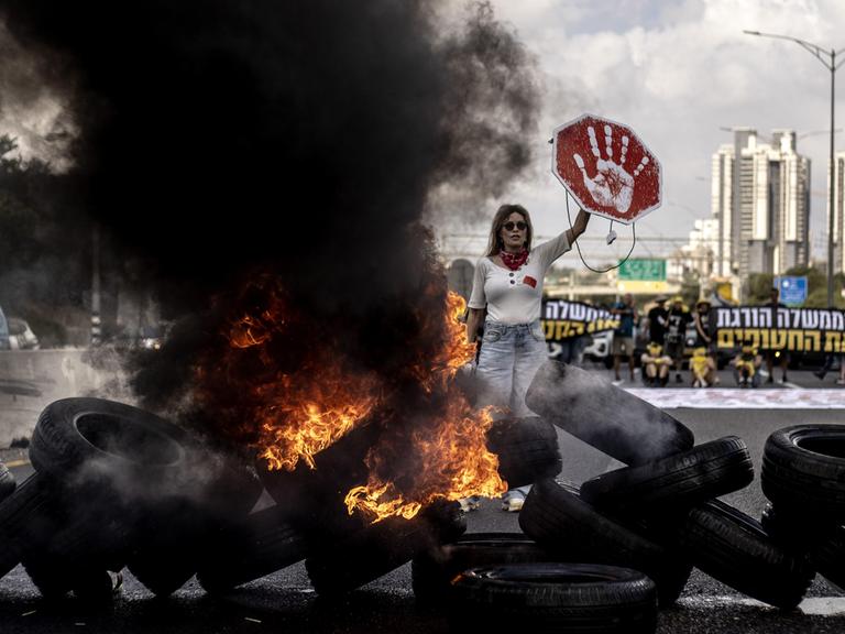 Demonstrantin in Tel Aviv (Israel) hält ein rotes Schild mit weißem Handabdruck in die Höhe; vor ihr liegt eine Barrikade aus brennenden Reifen, neben ihr steigt eine schwarze Rauchwolke in die Höhe (26.08.2025)