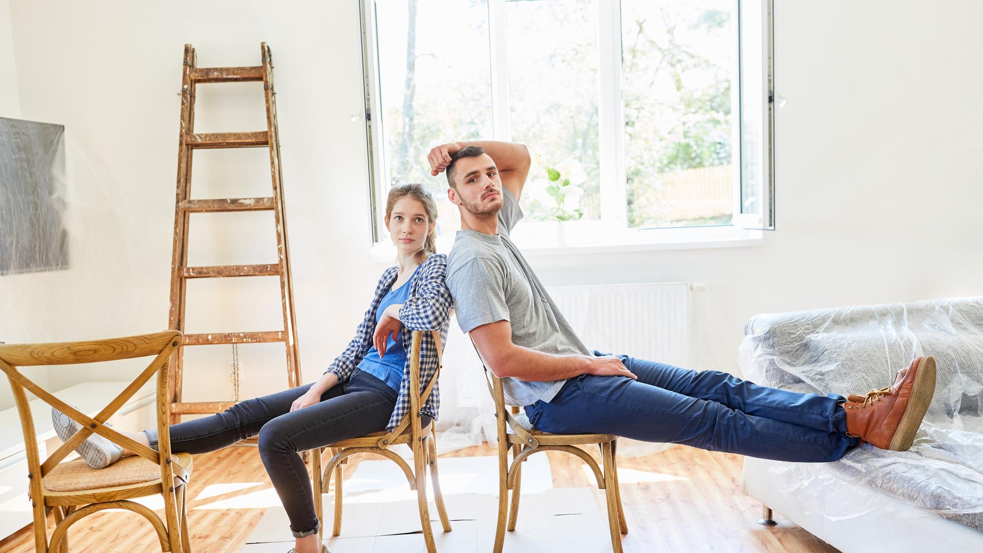 Junges Paar sitzt erschöpft und müde beim Einzug in eine neue Wohnung auf zwei Stühlen, die auf ausgefalteten Pappkartons stehen. Hinten an der Wand ist eine Leiter angelehnt. Das Sofa ist noch in Folie eingepackt. Junges Paar sitzt erschöpft und müde beim Einzug in eine neue Wohnung auf zwei Stühlen, die auf ausgefalteten Pappkartons stehen. Hinten an der Wand ist eine Leiter angelehnt. Das Sofa ist noch in Folie eingepackt.