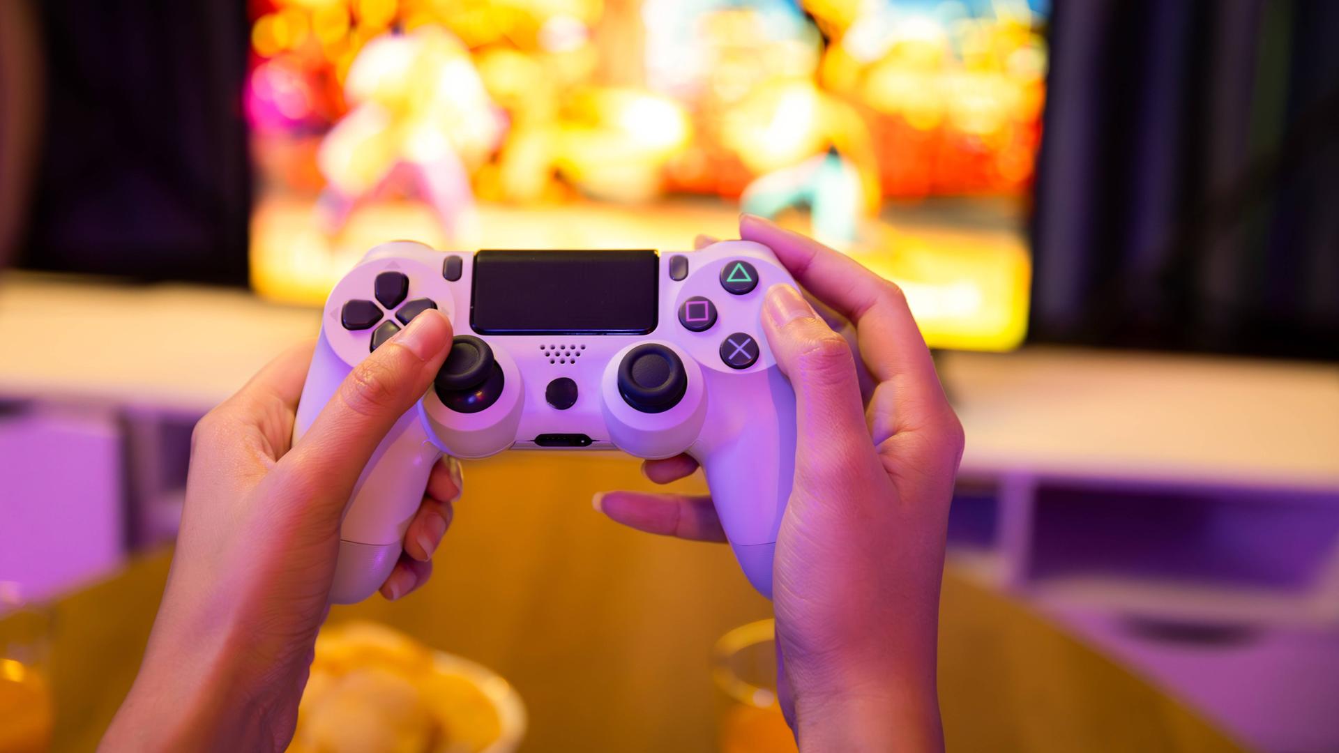Zwei Hände an einer Konsole und im Hintergrund läuft ein Computerspiel auf dem Bildschirm. Zwei Hände an einer Konsole und im Hintergrund läuft ein Computerspiel auf dem Bildschirm.