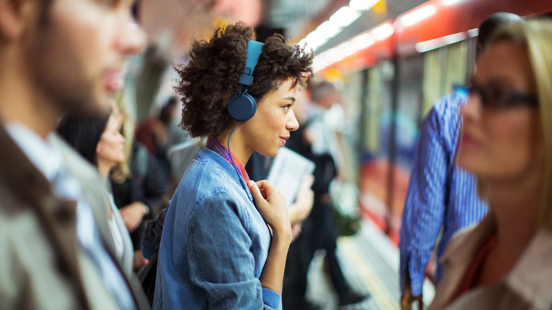 Eine Frau, die im Profil zu sehen ist, trägt blaue Kopfhörer und schaut offen und freundlich geradeaus. Um sie herum sind in der Unschärfe weitere Menschen zu sehen. Sie stehen an einem überfüllten Bahngleis.