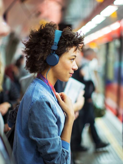 Eine Frau, die im Profil zu sehen ist, trägt blaue Kopfhörer und schaut offen und freundlich geradeaus. Um sie herum sind in der Unschärfe weitere Menschen zu sehen. Sie stehen an einem überfüllten Bahngleis.