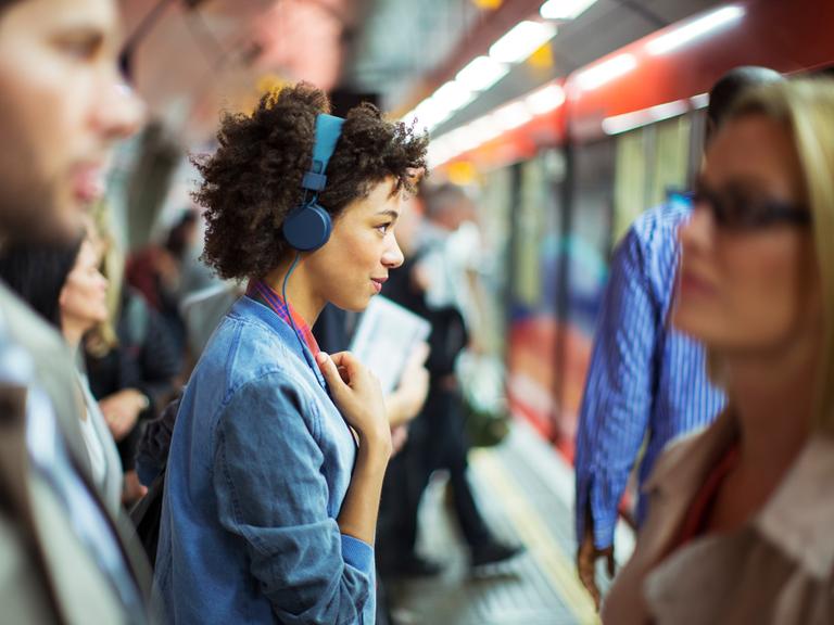 Eine Frau, die im Profil zu sehen ist, trägt blaue Kopfhörer und schaut offen und freundlich geradeaus. Um sie herum sind in der Unschärfe weitere Menschen zu sehen. Sie stehen an einem überfüllten Bahngleis.