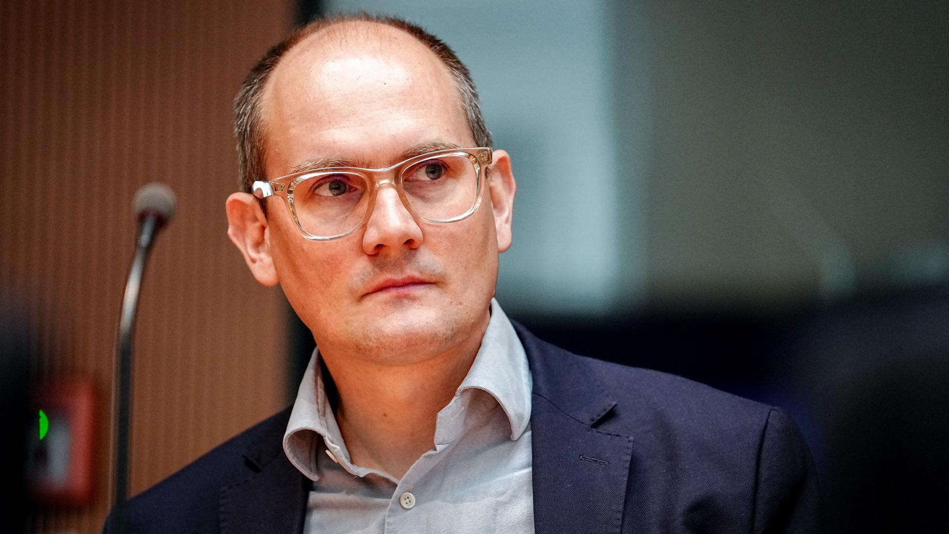 Der Grünen-Politiker Janosch Dahmen trägt ein graues Hemd, darüber ein dunkelblaues Sakko und ein transparentes Brillengestell. Er schaut seitlich in die Kamera. 
