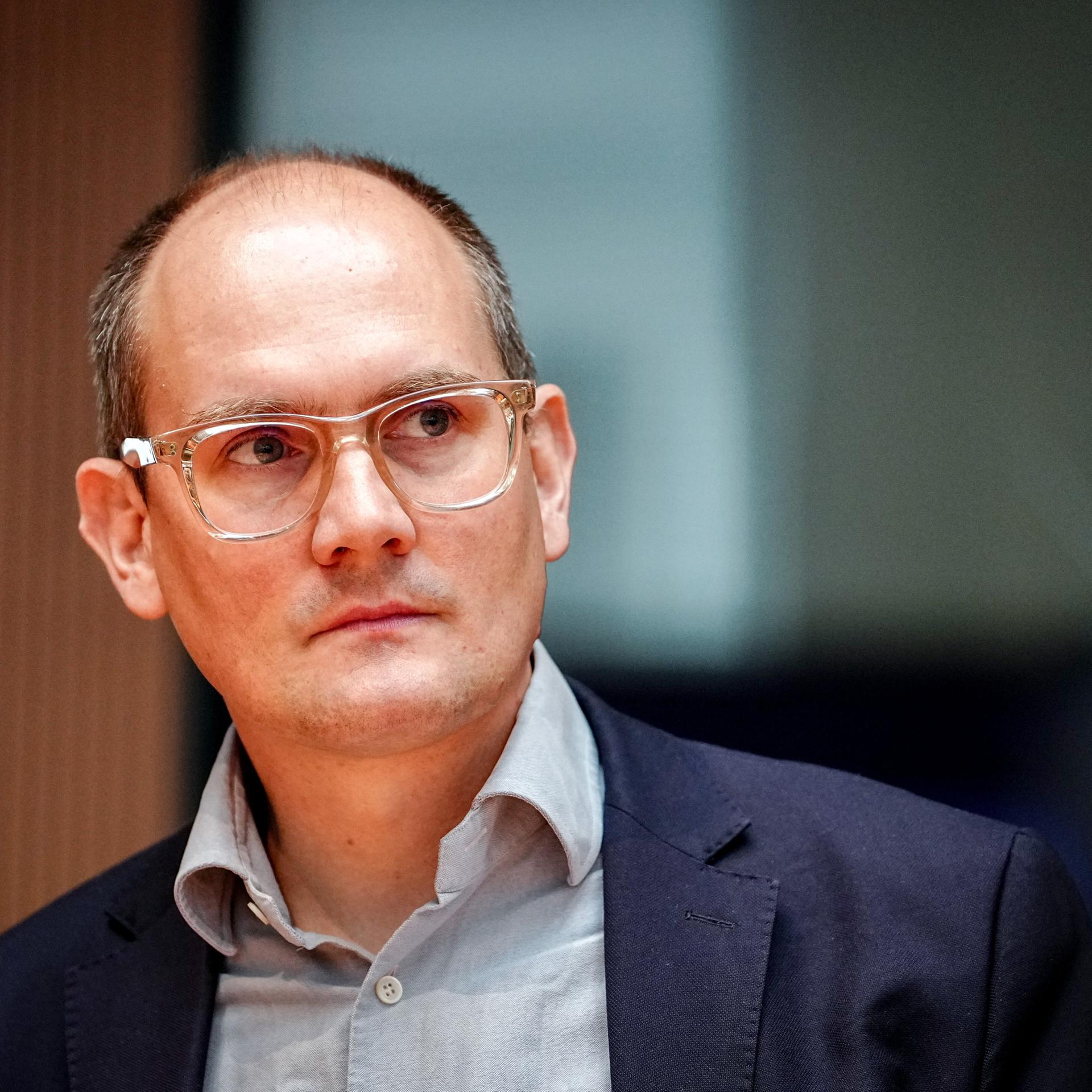 Der Grünen-Politiker Janosch Dahmen trägt ein graues Hemd, darüber ein dunkelblaues Sakko und ein transparentes Brillengestell. Er schaut seitlich in die Kamera. 
