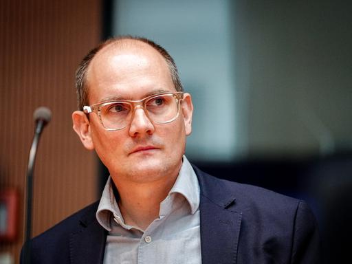 Der Grünen-Politiker Janosch Dahmen trägt ein graues Hemd, darüber ein dunkelblaues Sakko und ein transparentes Brillengestell. Er schaut seitlich in die Kamera. 
