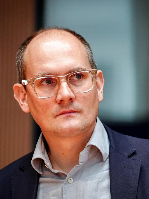 Der Grünen-Politiker Janosch Dahmen trägt ein graues Hemd, darüber ein dunkelblaues Sakko und ein transparentes Brillengestell. Er schaut seitlich in die Kamera. 
