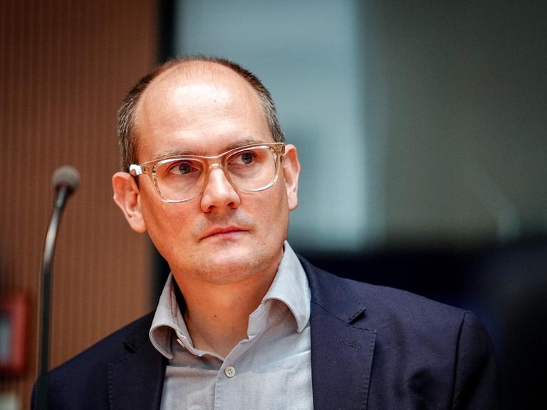 Der Grünen-Politiker Janosch Dahmen trägt ein graues Hemd, darüber ein dunkelblaues Sakko und ein transparentes Brillengestell. Er schaut seitlich in die Kamera. 
