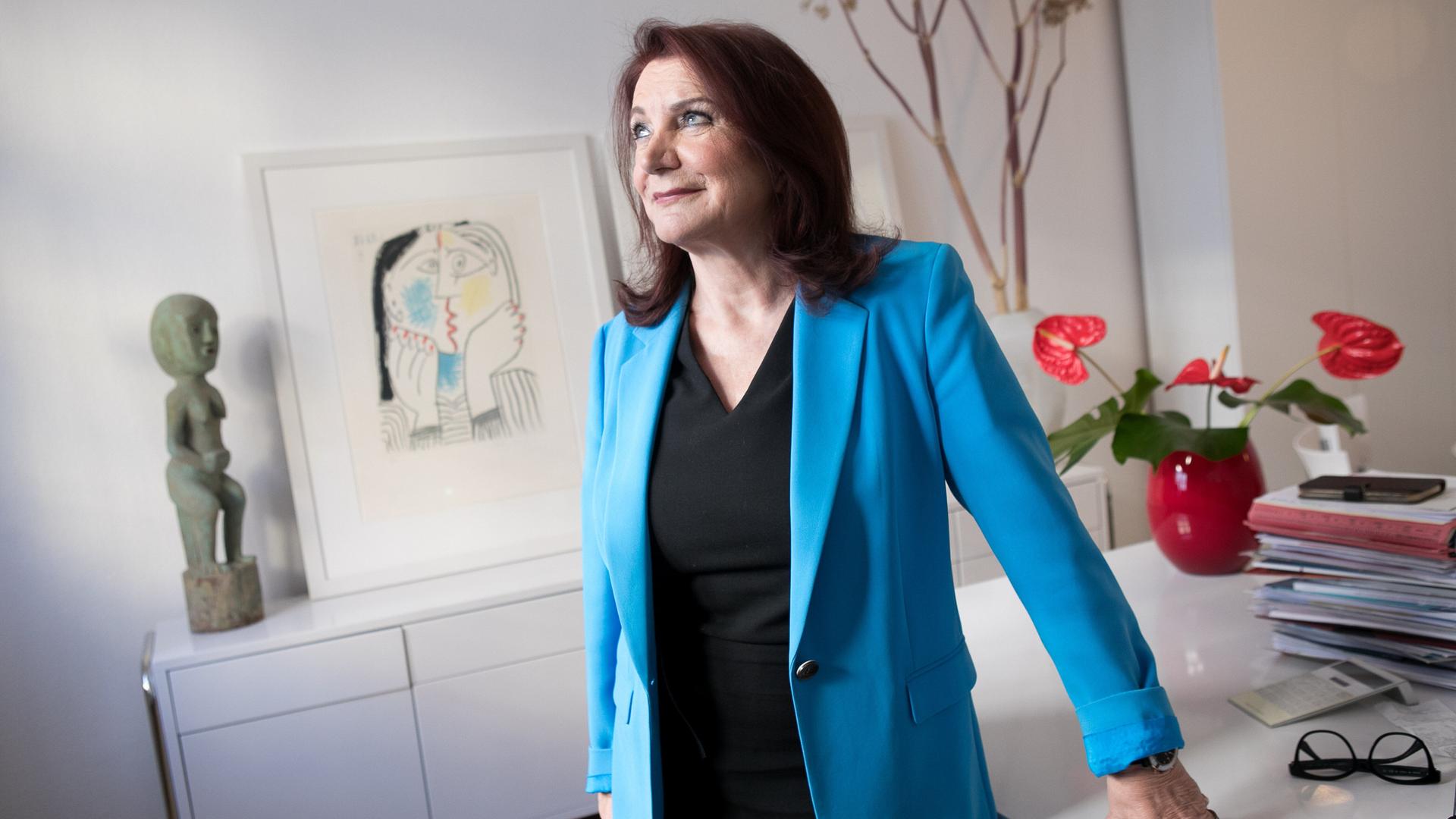 Eine dunkelhaarige Frau in blauem Blazer und schwarzem T-Shirt lehnt sich an einen Schreibtisch, auf dem unter anderem mehrere Akten liegen. Sie blickt zur Seite. Die Frau ist die Rechtsanwältin Renate Maltry, Eine dunkelhaarige Frau in blauem Blazer und schwarzem T-Shirt lehnt sich an einen Schreibtisch, auf dem unter anderem mehrere Akten liegen. Sie blickt zur Seite. Die Frau ist die Rechtsanwältin Renate Maltry,