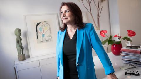 Eine dunkelhaarige Frau in blauem Blazer und schwarzem T-Shirt lehnt sich an einen Schreibtisch, auf dem unter anderem mehrere Akten liegen. Sie blickt zur Seite. Die Frau ist die Rechtsanwältin Renate Maltry,