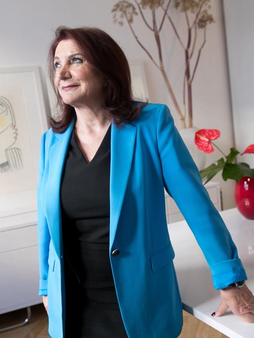 Eine dunkelhaarige Frau in blauem Blazer und schwarzem T-Shirt lehnt sich an einen Schreibtisch, auf dem unter anderem mehrere Akten liegen. Sie blickt zur Seite. Die Frau ist die Rechtsanwältin Renate Maltry,