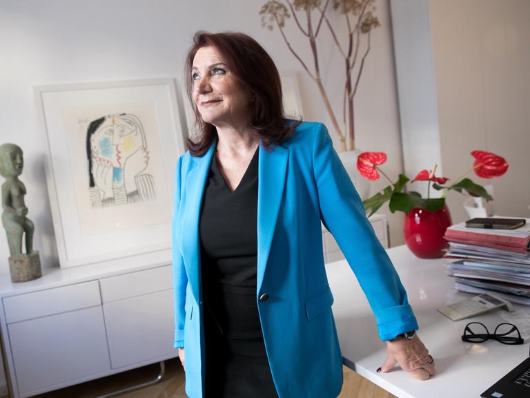 Eine dunkelhaarige Frau in blauem Blazer und schwarzem T-Shirt lehnt sich an einen Schreibtisch, auf dem unter anderem mehrere Akten liegen. Sie blickt zur Seite. Die Frau ist die Rechtsanwältin Renate Maltry,