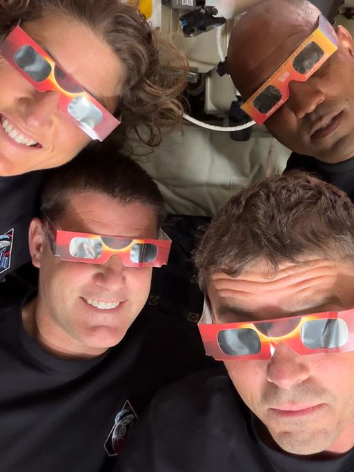 Vier Mitglieder der Artemis‑II‑Besatzung liegen mit den Köpfen eng beieinander in einer Raumfahrzeug‑Umgebung und tragen rot‑orange Sonnenfinsternisbrillen.