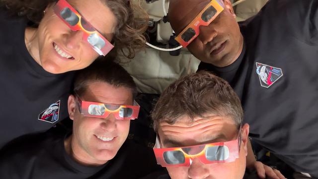 Vier Mitglieder der Artemis‑II‑Besatzung liegen mit den Köpfen eng beieinander in einer Raumfahrzeug‑Umgebung und tragen rot‑orange Sonnenfinsternisbrillen.