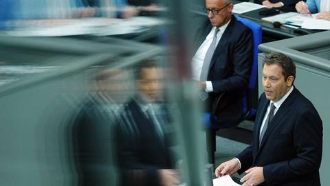 Bundesfinanzminister Lars Klingbeil (r., SPD) spricht am Rednerpult im Bundestag, im Bild spiegelt er sich dabei unscharf in einer Glasscheibe. Im Hintergrund sitzt Bundeskanzler Friedrich Merz (CDU) auf der Regierungsbank
