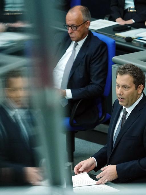 Bundesfinanzminister Lars Klingbeil (r., SPD) spricht am Rednerpult im Bundestag, im Bild spiegelt er sich dabei unscharf in einer Glasscheibe. Im Hintergrund sitzt Bundeskanzler Friedrich Merz (CDU) auf der Regierungsbank