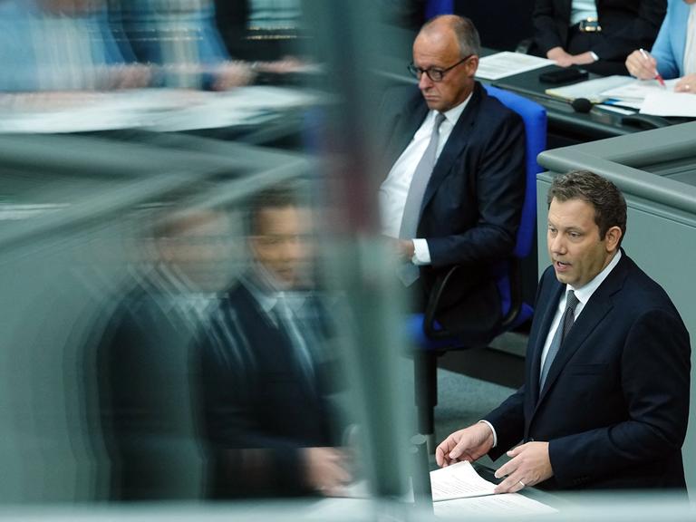 Bundesfinanzminister Lars Klingbeil (r., SPD) spricht am Rednerpult im Bundestag, im Bild spiegelt er sich dabei unscharf in einer Glasscheibe. Im Hintergrund sitzt Bundeskanzler Friedrich Merz (CDU) auf der Regierungsbank