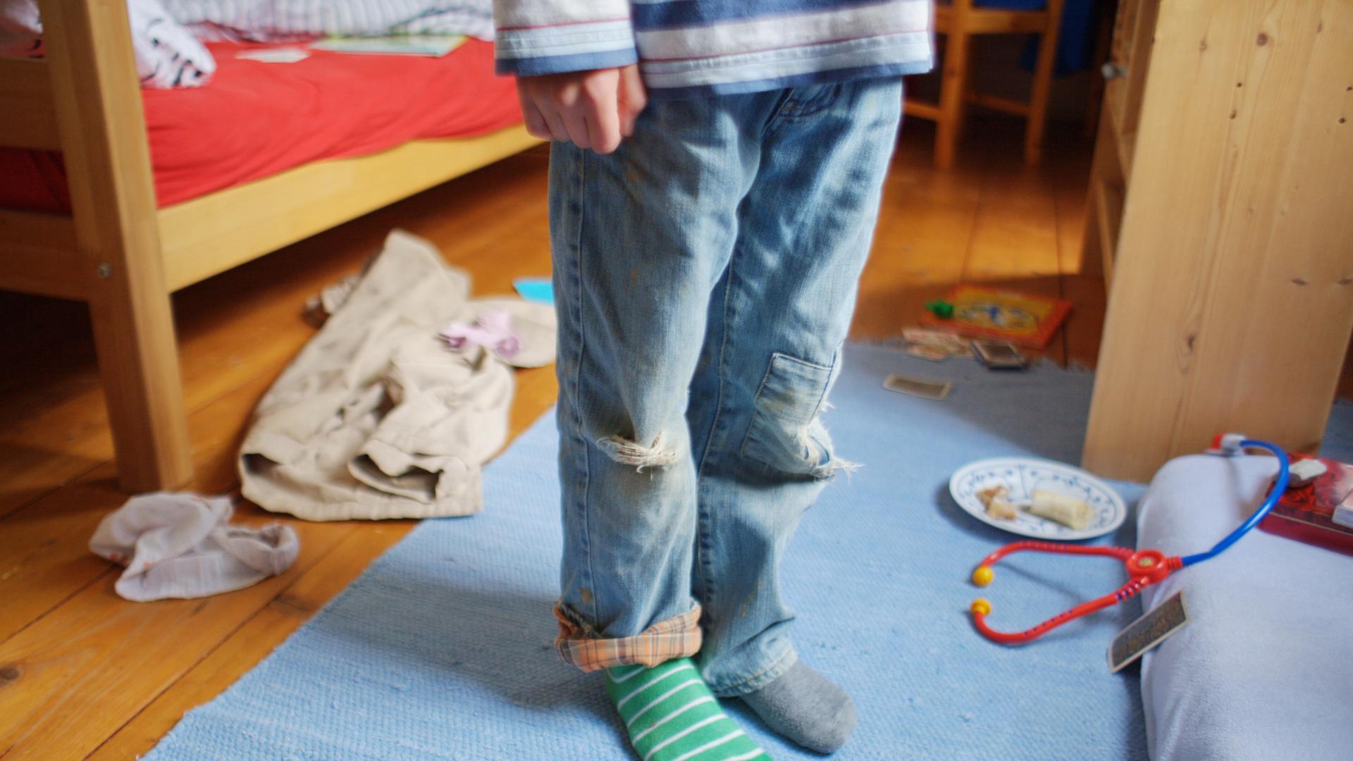 Ein Kind steht mit schmutziger Kleidung, zerrissenen Hosen und verschiedenen Strümpfen in einem unaufgräumten Zimmer.