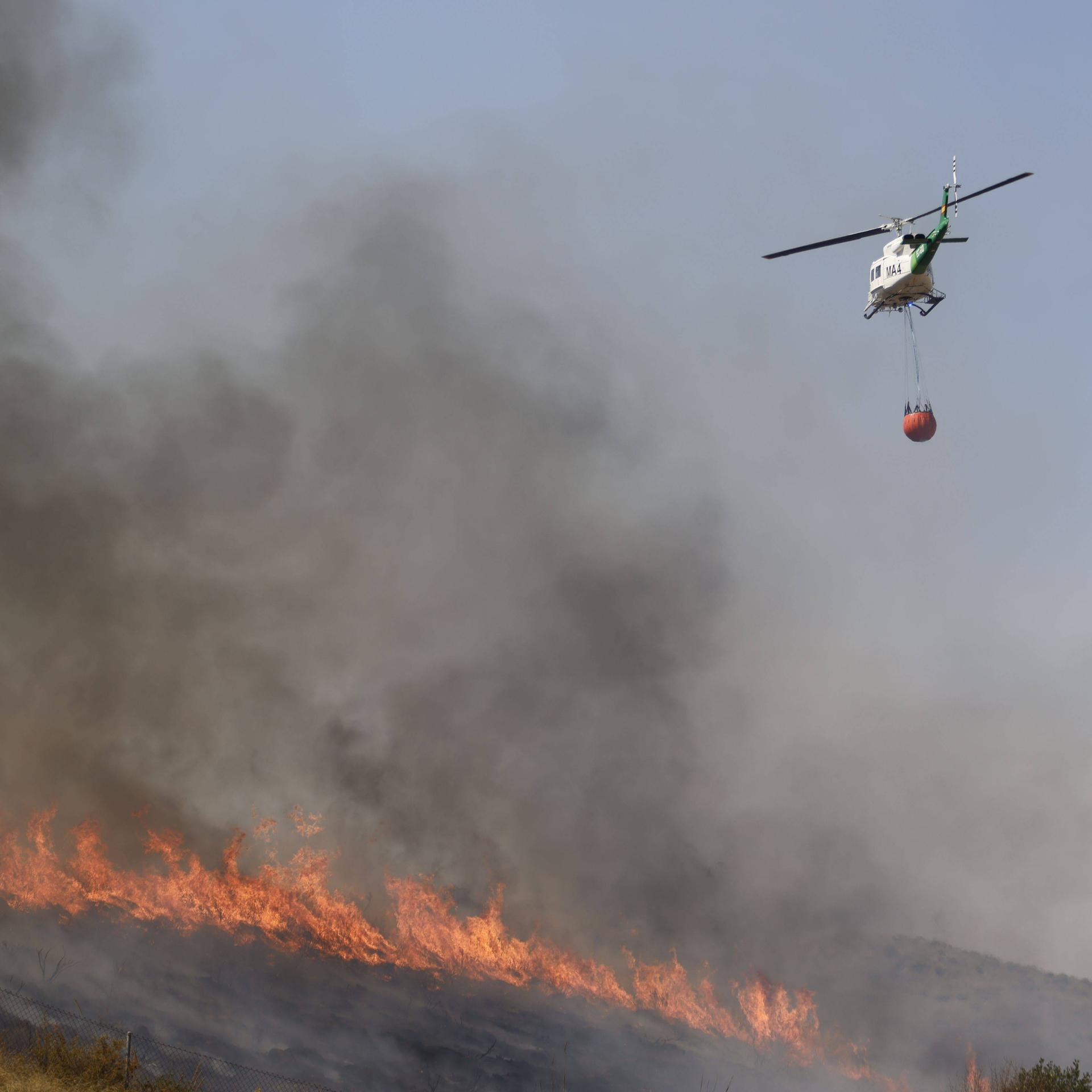 Ein Löschhubschrauber fliegt über einem brennenden Waldgebiet.