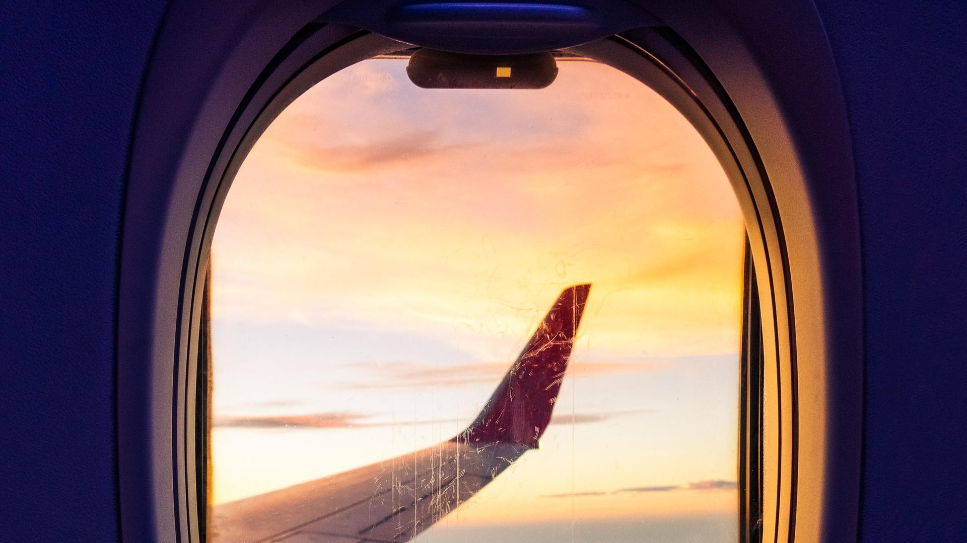 Blick durch ein Flugzeugfenster mit orangefarbenen Wolken bei Sonnenuntergang.