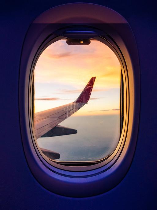 Blick durch ein Flugzeugfenster mit orangefarbenen Wolken bei Sonnenuntergang.