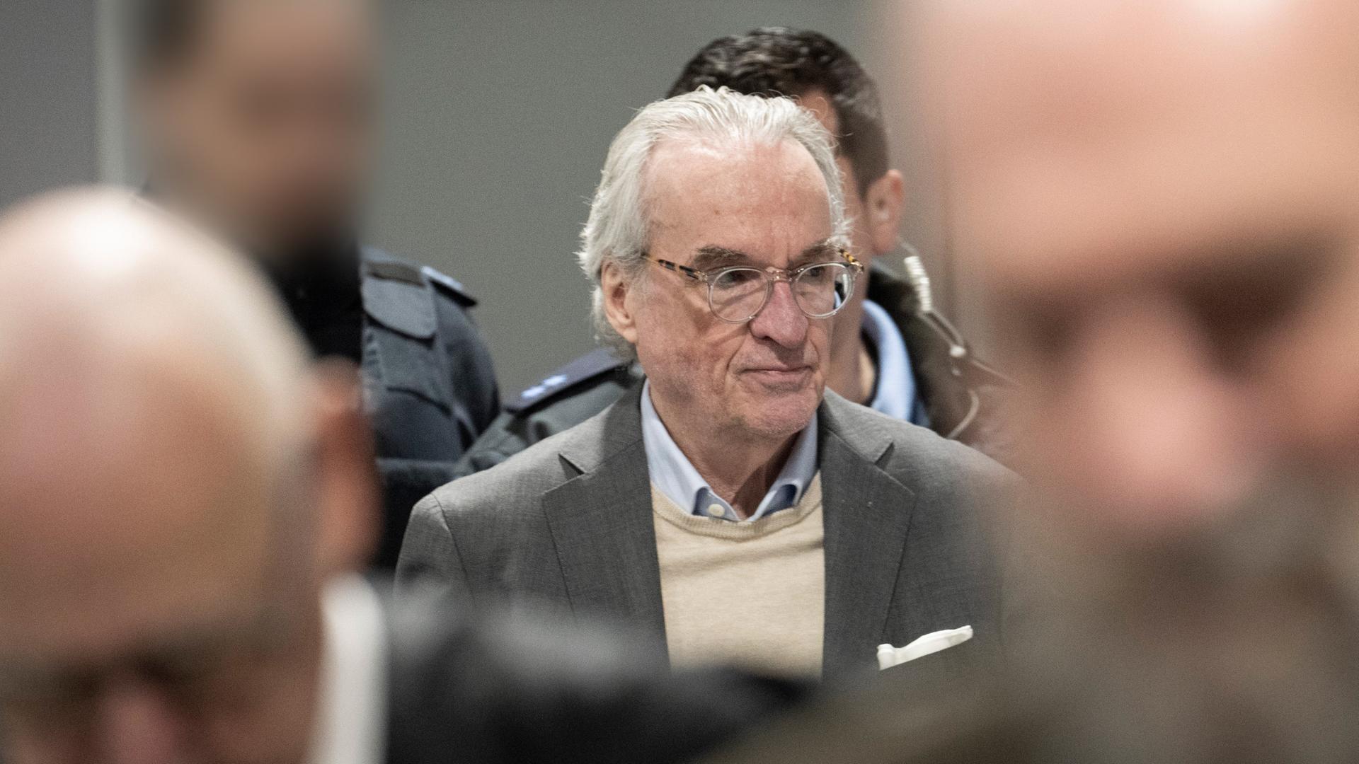 Nahaufnahme von Reuß vor zwei verpixelten Justizbeamten. Davor unscharf die Köpfe zweier weiterer Personen. Nahaufnahme von Reuß vor zwei verpixelten Justizbeamten. Davor unscharf die Köpfe zweier weiterer Personen.