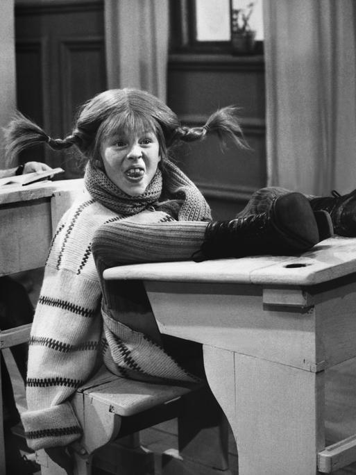 Inger Nilsson als Pippi Langstrumpf in einer Filmszene, in der Pippi ihre Füße auf die Schulbank legt und frech schaut