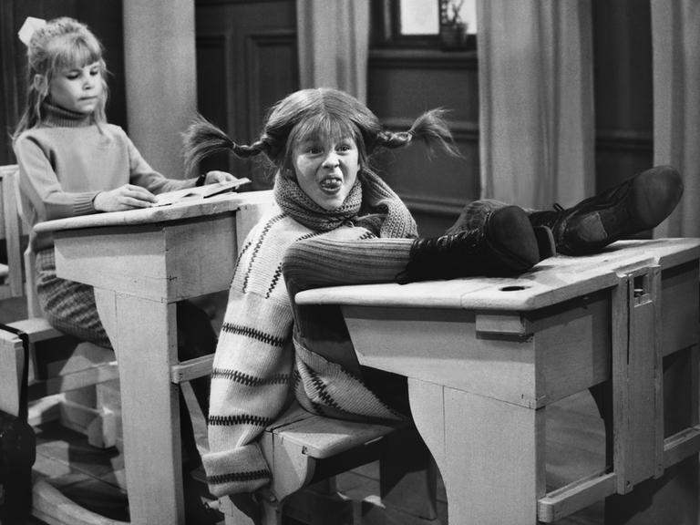 Inger Nilsson als Pippi Langstrumpf in einer Filmszene, in der Pippi ihre Füße auf die Schulbank legt und frech schaut