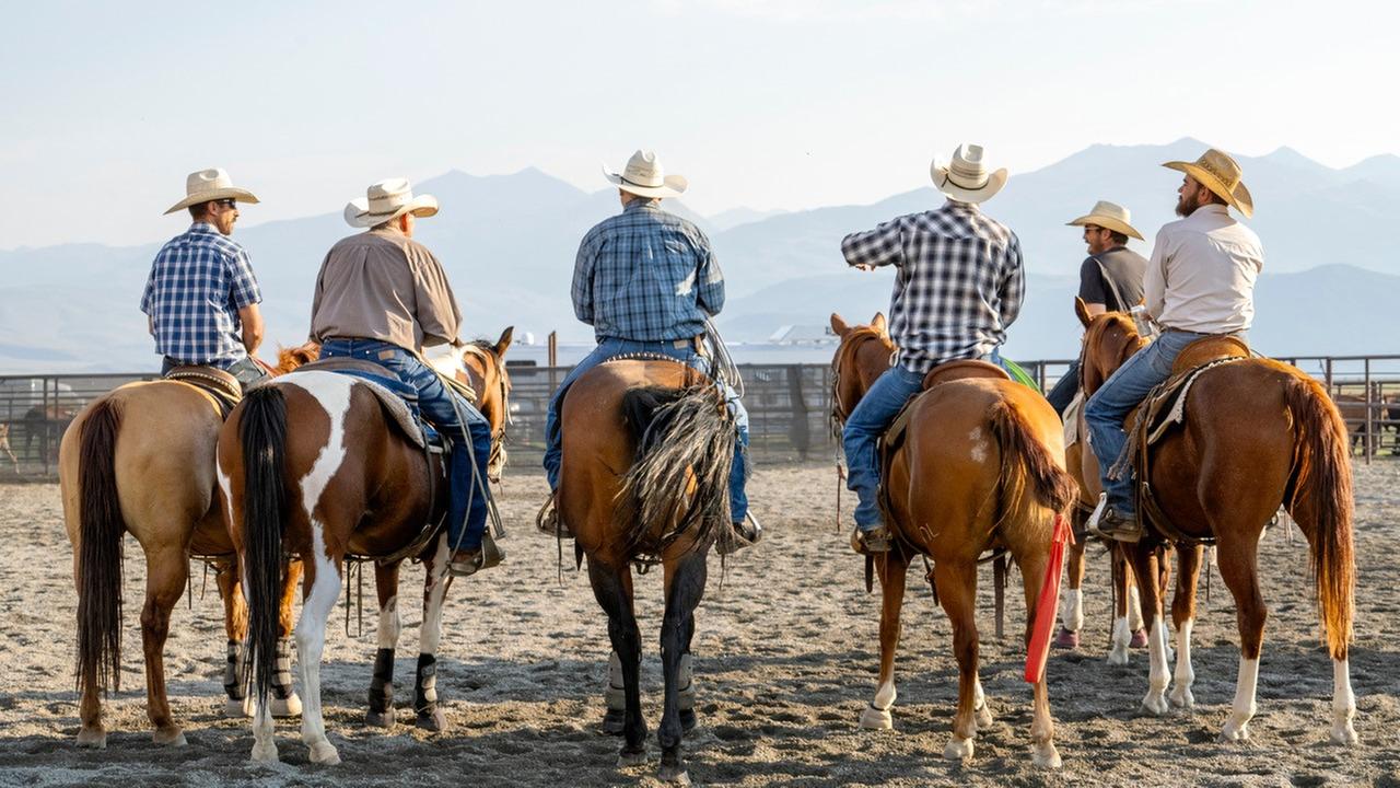 Mehrere Personen mit Cowboyhüten auf Pferden sitzend, auf sandigem Untergrund nebeneinander aufgereiht und vor einer Bergkulisse.