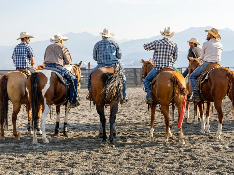 Mehrere Personen mit Cowboyhüten auf Pferden sitzend, auf sandigem Untergrund nebeneinander aufgereiht und vor einer Bergkulisse.