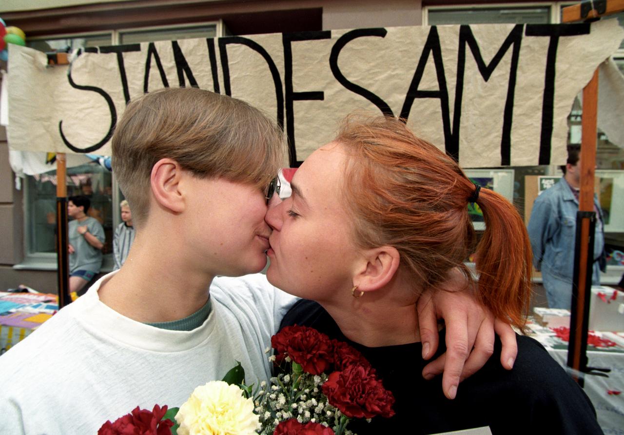 Zwei Frauen küssen sich. Dahinter ein Pappschild mit der Aufschrift Standesamt.