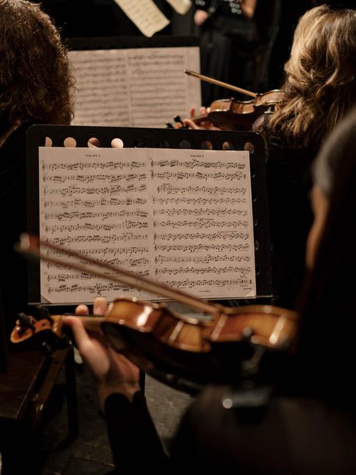 Blick über verschiedene Geigenpulte in einem Orchester, das gerade spielt.