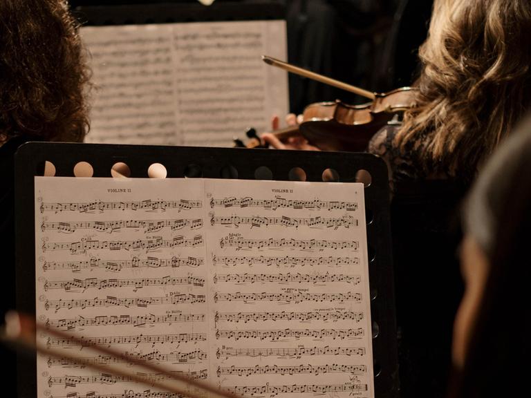 Blick über verschiedene Geigenpulte in einem Orchester, das gerade spielt.