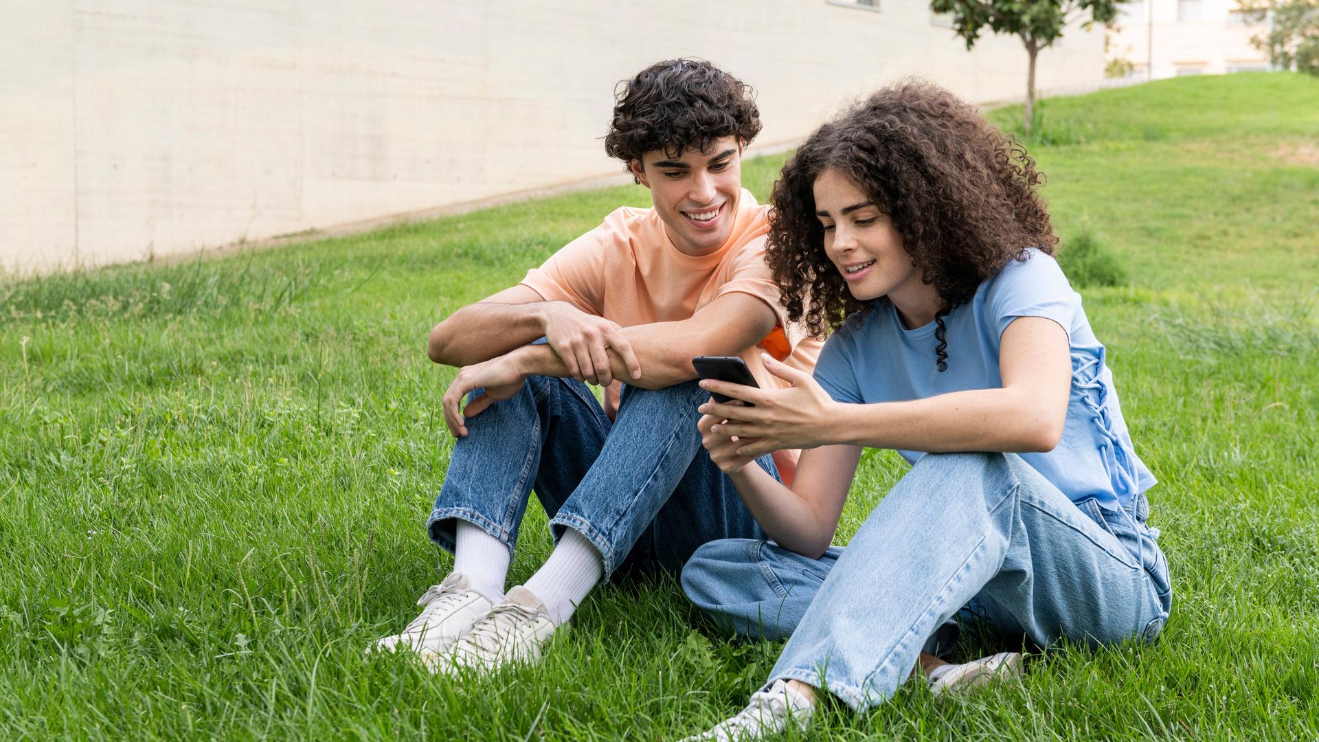Ein junges paar sitzt lächelnd in der Wiese und schaut auf ein Smartphone.