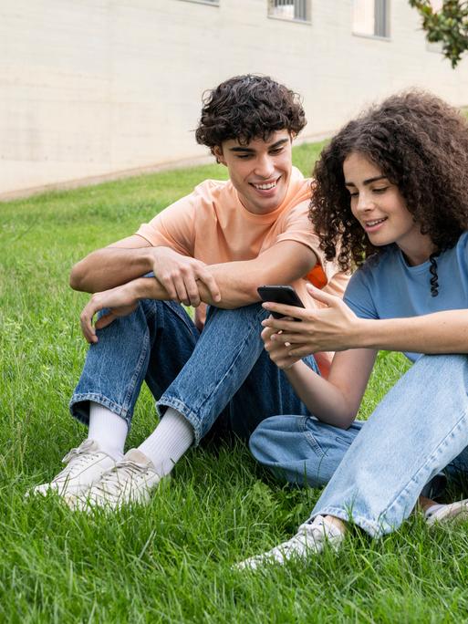 Ein junges paar sitzt lächelnd in der Wiese und schaut auf ein Smartphone.