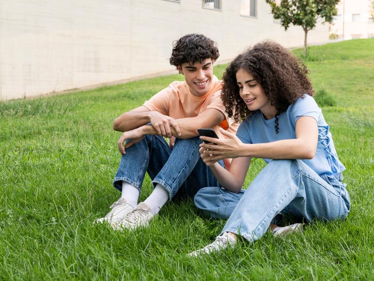 Ein junges paar sitzt lächelnd in der Wiese und schaut auf ein Smartphone.