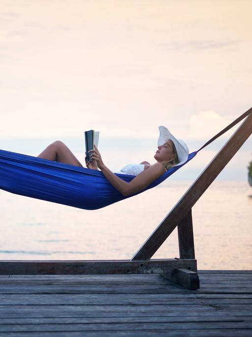 Eine Frau liest in einer Hängematte auf einem Steg ein Buch. Im Hintergrund ist das Wasser zu sehen.