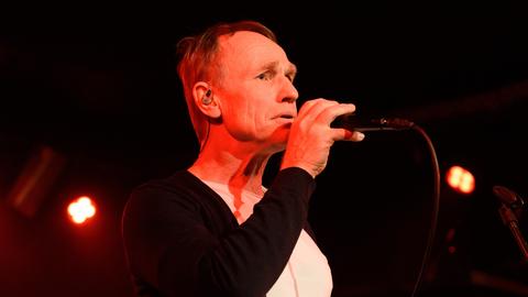 Norbert Leisegang von "Keimzeit" bei einem Konzert in München im April 2024 auf der Bühne am Mikrofon vor schwarzem Hintergrund. Er trägt ein weisses Hemd und eine schwarze Jacke.