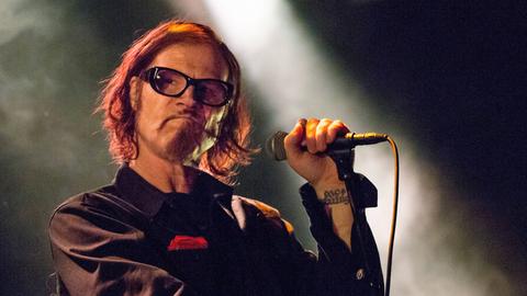 Mark Lanegan, mit halblangen Haaren, Bart und einer markanten Brille, bei einem Auftriff auf der Bühne. Der Strahl des Scheinwerfers trifft seine Hand, die das Mikrofon hält.